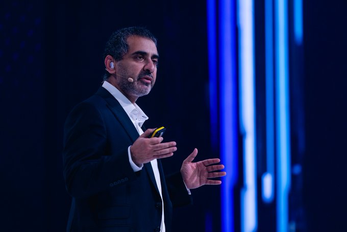 A man in a dark suit with a headset microphone speaks on stage against a vibrant blue illuminated backdrop, holding a presentation clicker. The scene captures the moment referenced in the post, showing @balajis delivering his keynote on the OKX Mainstage about the real-world impact of crypto communities.