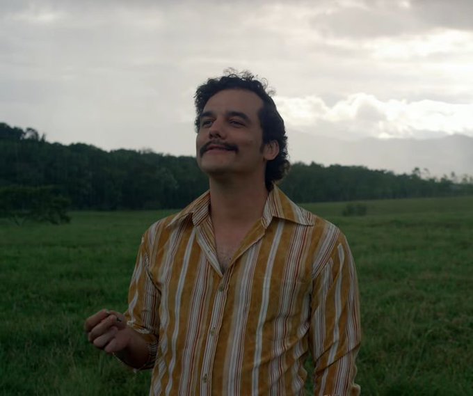 A man with a mustache wearing a striped orange and white shirt stands in a grassy field under a cloudy sky, smiling softly with his eyes closed.
