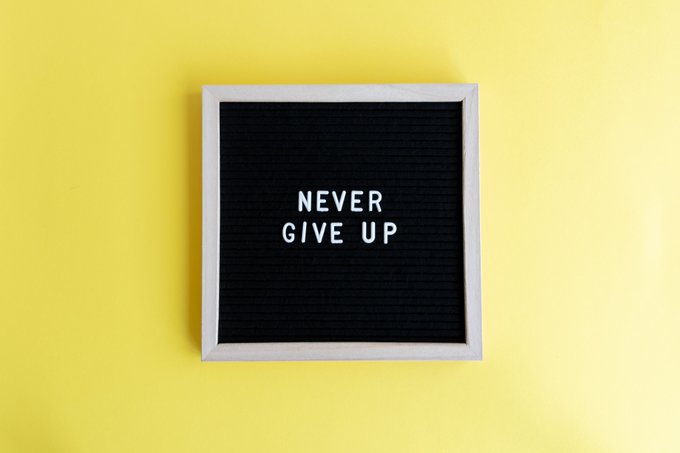 A white-framed black felt letter board on a bright yellow wall displays the text 'NEVER GIVE UP' in white letters.