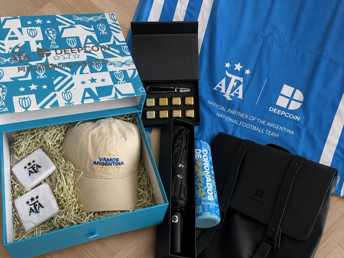 A gift box from DeepCoin, the official partner of the Argentina national football team, containing a beige "Vamos Argentina" cap, AFA-branded cufflinks, gold coins, a black umbrella, a "Dogomados" tumbler, and a leather bag, displayed alongside a blue AFA x DeepCoin jersey. The image showcases the branded merchandise received as a sponsorship gift, highlighting DeepCoin's collaboration with the Argentine Football Association (AFA).