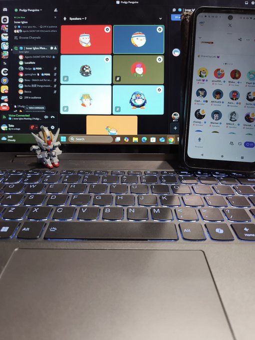 A laptop displaying a Discord video call with multiple participants sits next to a phone showing what appears to be a social media feed; a small Gundam figurine is placed on the laptop keyboard.