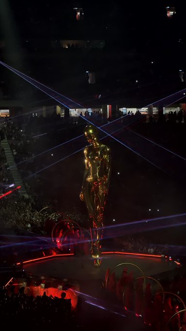 A golden humanoid statue stands illuminated on stage at an award ceremony, surrounded by dramatic laser beams and red lighting.