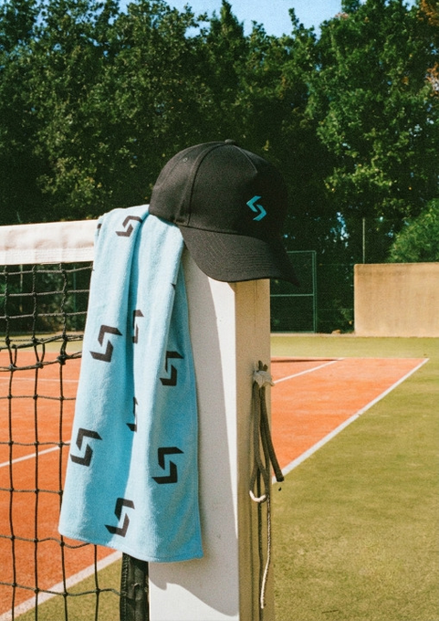 A black baseball cap with a blue logo rests on a tennis net post alongside a light blue towel patterned with matching logos, set against an outdoor clay court.