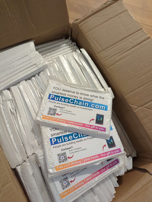 A cardboard box filled with stacks of promotional cards for PulseChain, reading "YOU deserve to know what the smartest money is doing!" and "Free LCD Writing Tablet Inside - Our gift to you," featuring a QR code and the tagline "Curious? Scan To Explore. It only takes a moment."