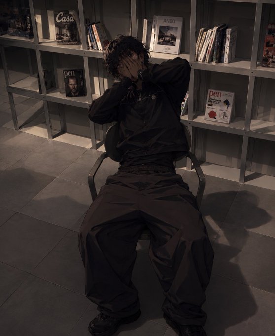 A person dressed in all black sits in a chair in a dimly lit room lined with bookshelves, covering their face with their hands.