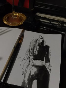 A black and white ink fashion sketch of a woman on white paper, sitting next to a gold pen and an ornate golden candlestick on a dark surface.
