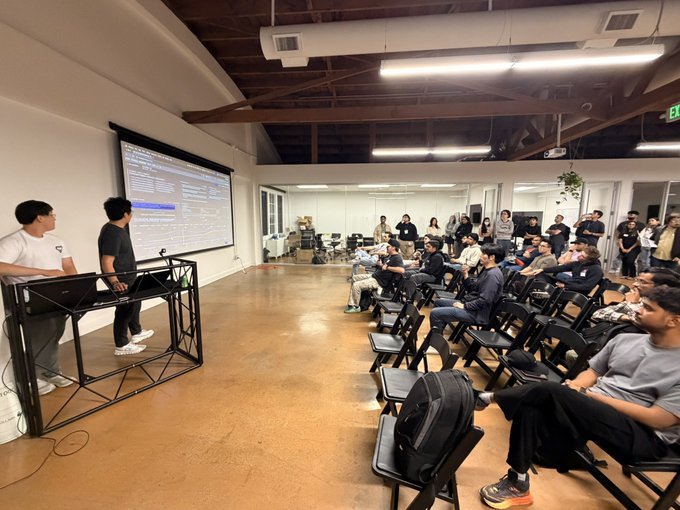 The image shows a group of people sitting in chairs, with some standing behind a podium or stage. The room appears to be a conference or meeting space where an event is taking place. There are several individuals present, including two men standing at the front of the room and many others seated around them. A large screen can be seen on one side of the room, possibly displaying information relevant to the ongoing discussion.

The people in the image seem engaged in a conversation or presentation, with some holding backpacks nearby. The overall atmosphere appears to be professional and focused, as everyone seems to be actively participating in the event.