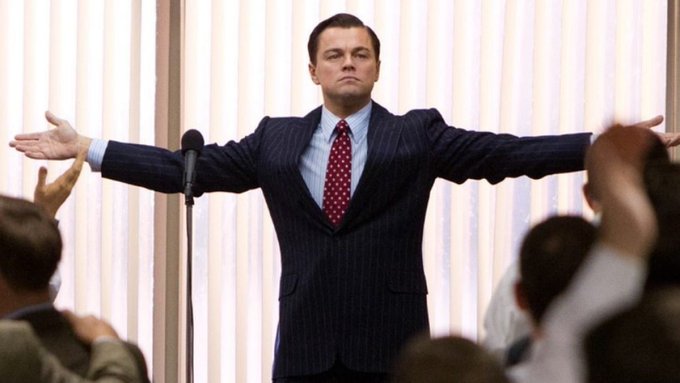 A still image of Leonardo DiCaprio in a pinstripe suit with a red polka-dot tie, standing with his arms outstretched while addressing a crowd from a stage with a microphone.