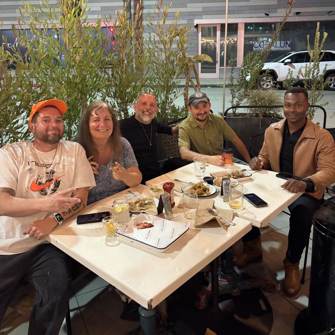 A group of five people smiling and posing together at a restaurant table with food and drinks.