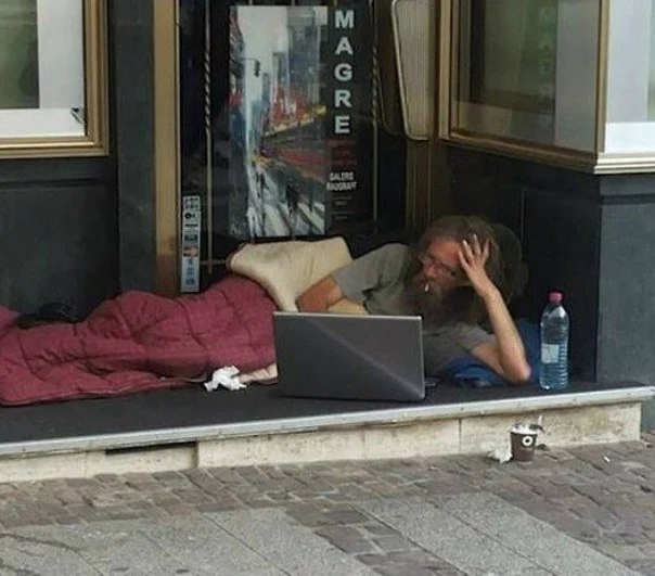 The image shows a man lying down on the ground, using his laptop computer while surrounded by various objects such as a bottle of water, a cup, and some trash nearby. The scene appears to be set outdoors with a building visible behind him. A caption in the image reads "Me buying another dip." This phrase is likely an inside joke or reference to someone else's recent actions.