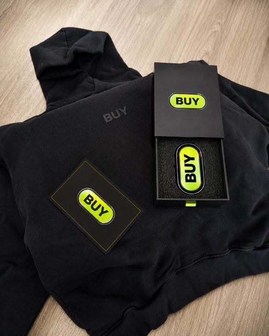 A merchandise photo showing a black hoodie and a presentation box containing an enamel pin, all branded with a neon green 'BUY' logo, laid out on a wooden surface.