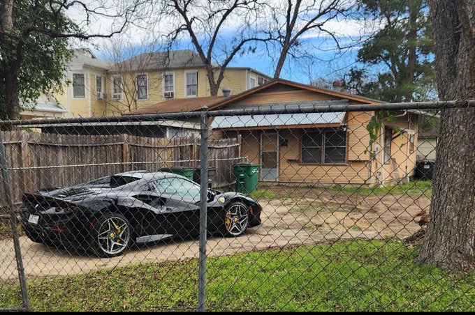 A damaged black sports car (appears to be a Ferrari) parked behind a chain-link fence in the driveway of a modest beige house with green trash bins.