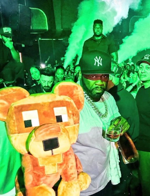 A nighttime party scene bathed in green light and smoke, featuring a man in a Yankees cap and chain holding a drink alongside a large orange pixelated bear plush, with a bottle of liquor and a crowd in the background.