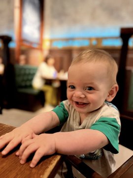 A happy toddler with light brown hair in a green and white shirt smiling and leaning on a wooden high chair in what appears to be a restaurant setting.