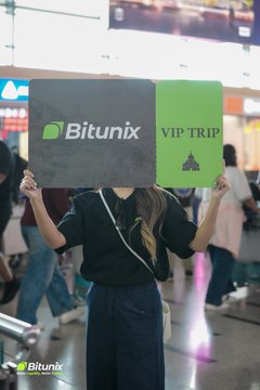 A woman holds a "Bitunix X VIP TRIP" sign in what appears to be an airport or transit hub, marking the arrival phase of the Ultra Arena tournament weekend. The branded sign connects to the post's description of Day 1 arrivals as traders gathered before the competition commenced.