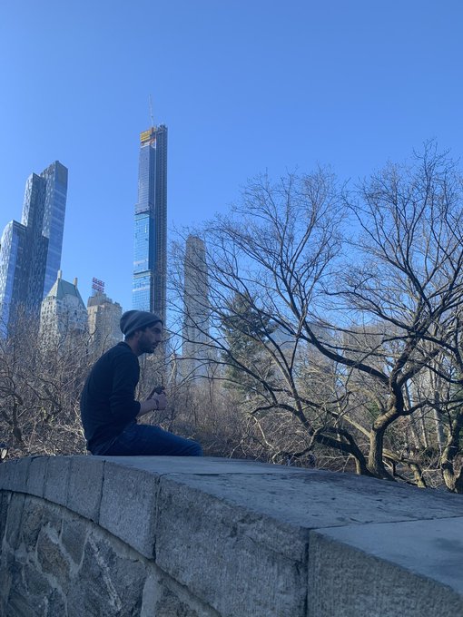 A man wearing a beanie and sunglasses sitting on a stone wall overlooking a city skyline with tall skyscrapers and bare trees.