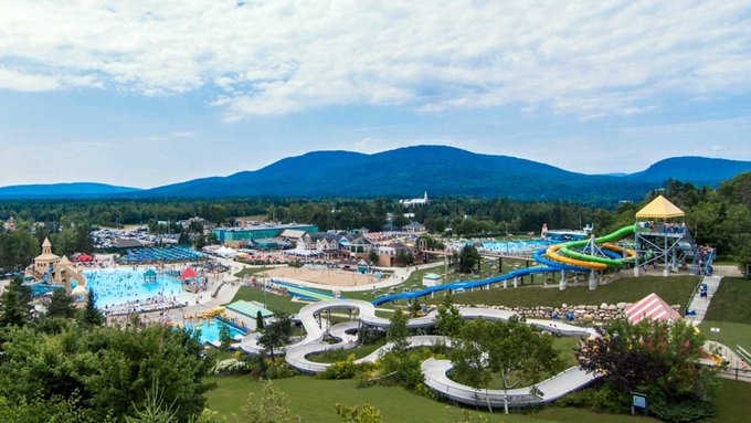 An aerial view of a large water park featuring blue pools and multiple water slides, set against a backdrop of green fields, trees, and rolling mountains under a partly cloudy sky.