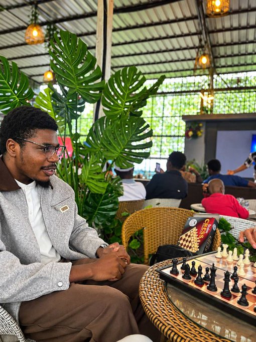 A young man in a gray jacket and glasses sits in a lush outdoor cafe setting with tropical plants and hanging lights, playing chess with an opponent whose hand is visible moving pieces.