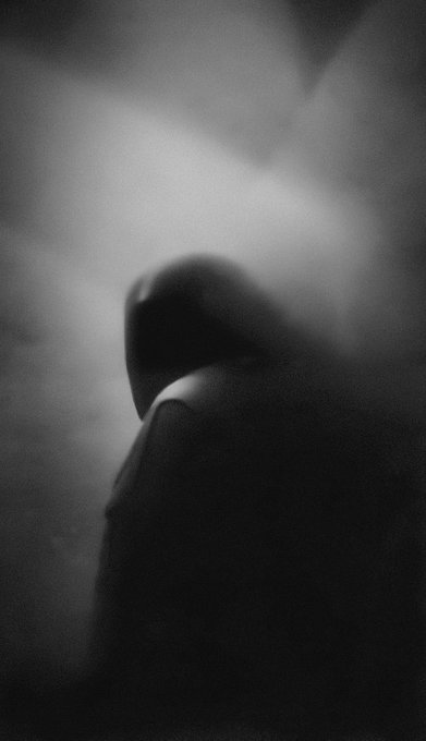 A moody, black-and-white artistic photograph of a hooded figure emerging from shadowy mist, with only the silhouette visible.