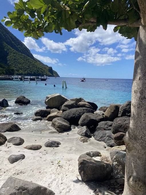 A tropical beach scene features white sand, large dark rocks, and clear turquoise water under a blue sky with scattered clouds, framed by green tree leaves in the foreground and a forested mountain on the left.