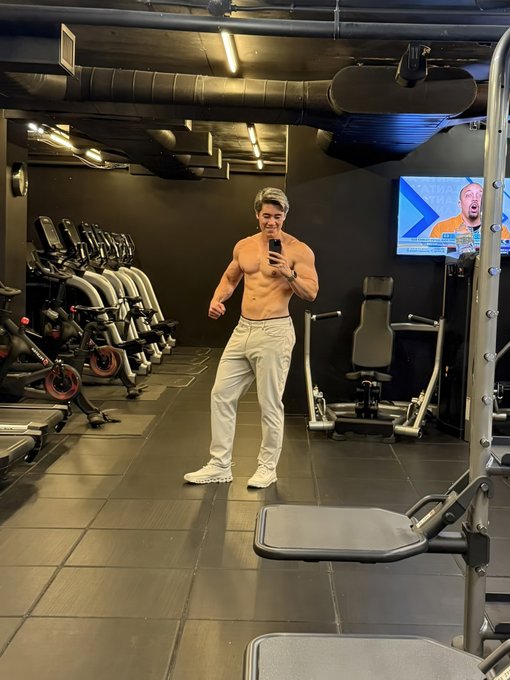 A shirtless, muscular man takes a mirror selfie in a gym, wearing white pants and white sneakers, with exercise equipment and a TV visible in the background.