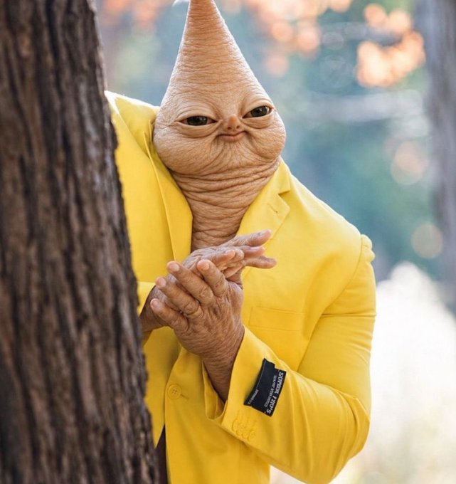 A creature wearing a yellow suit peeks out from behind a tree, clasping its hands together.