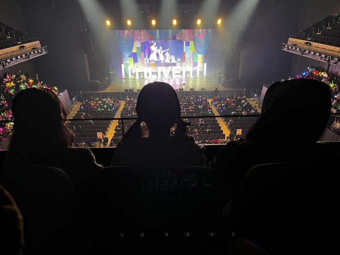 The image shows a group of people sitting in front of a stage at a theater or concert venue. They are watching a performance with bright lights illuminating the area, creating an immersive experience for them. The photo captures their silhouettes against the backdrop of the stage, emphasizing the atmosphere and excitement of the event.