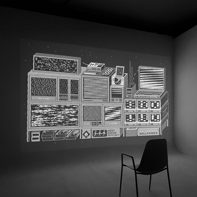 A monochrome pixel-art projection of stacked retro computer monitors and servers displaying various patterns and static illuminates a darkened room with a lone chair facing it. Text on the displays reads "US 26," "88:90," "INDEX ARCHIVE SKYLIGHT," and "WIRED WALLPAPERS," connecting to the "DREAM MACHINES" SuperRare fundraiser auction for Rhizome by @0xassembly.