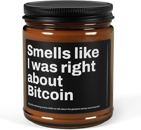 A brown jar candle with a black label reading 'Smells like I was right about Bitcoin' with smaller text about intoxicating aroma while talking about the greatest money ever invented.