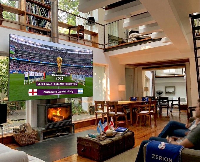 A large TV in a loft-style living room displays "FIFA CUP 2026 SEMI FINALS ENGLAND v ARGENTINA" with "Zerion World Cup Watch Party" overlaid, featuring small England and Argentina flags on the coffee table. Two friends relax on the couch watching the match, fitting the post about hosting an Argentina vs England watch party with Zerion in Lisbon.