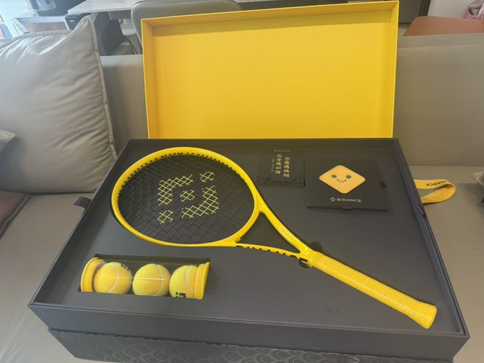 The image features a tennis racket, three tennis balls, and a box containing these items. The tennis racket has yellow strings and a black handle. There are also two other tennis rackets placed inside the box along with some tennis balls. The box is open, revealing its contents.