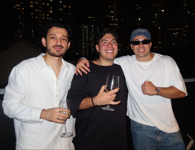 Three men pose together at a nighttime rooftop event, with two holding wine glasses and the man on the right wearing a cap reading "PEDROVI" along with sunglasses and a watch. The city skyline of Singapore glows in the background, fitting the Token2049 Singapore October 2026 crypto conference setting.