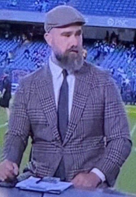 A man with a beard wearing a flat cap, white shirt, tie, and grey checkered blazer, sitting at a desk with a notebook in what appears to be a sports stadium setting.