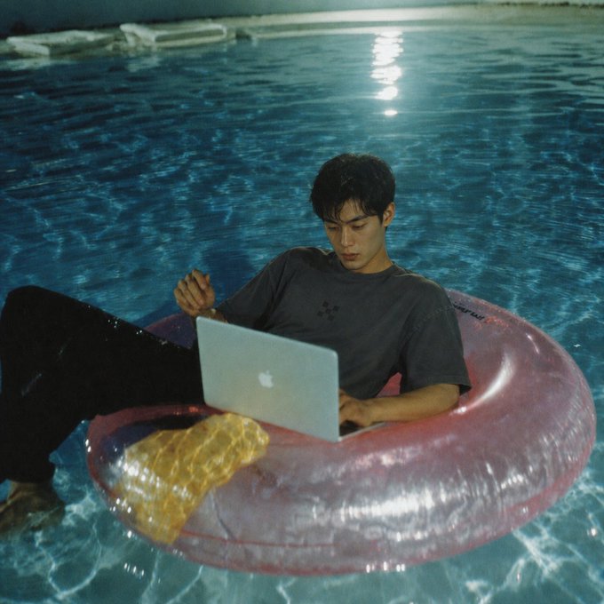 A slightly grainy, cinematic photo of a young man floating in a swimming pool on a pink inner tube, working intently on a silver Apple laptop while holding a pen.