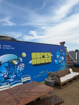 A vibrant rooftop deck at Arbitrum's Founder House in London features a colorful mural reading "POWERED BY ARBITRUM" and "SUPERHOUSE LONDON" alongside cartoon characters, with wooden picnic tables and blue sky overhead. The relaxed outdoor setting complements the post's description of a productive first day of workshops at the event, where the author built a new project on Robinhood Chain for tokenized stock positions.