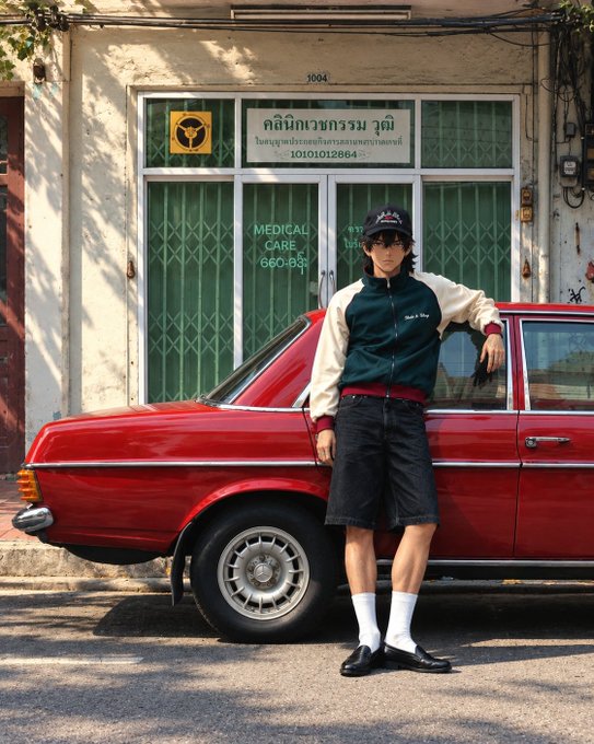 A fashion-style photo of a person in a green and cream varsity jacket, dark shorts, white socks, loafers, and a black cap, leaning against a red vintage car parked outside a Thai medical clinic.
