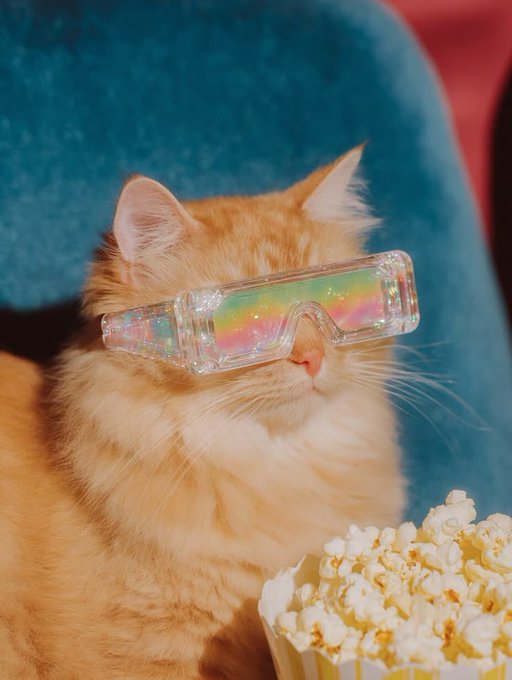 A fluffy orange cat wearing iridescent rainbow-colored 3D glasses sits in front of a blue chair next to a bucket of popcorn.