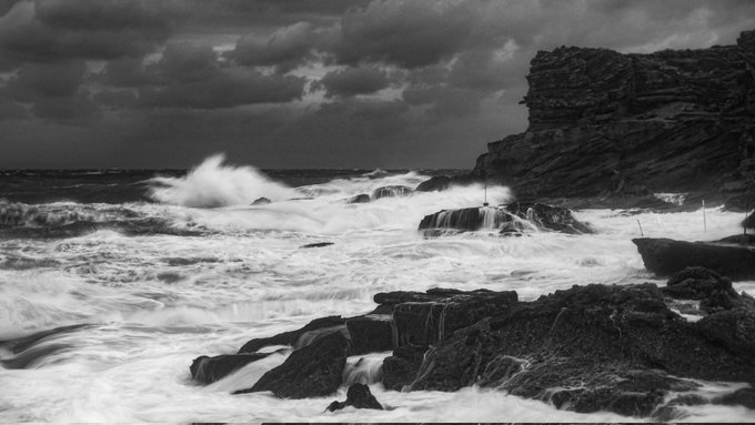 Black-and-white long-exposure photograph of turbulent ocean waves crashing against rocky cliffs under a dramatic, cloud-filled sky.