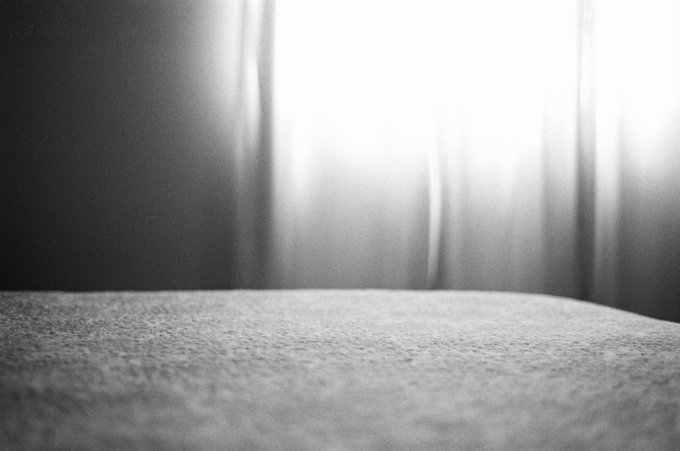 A black and white close-up photo of a textured surface, possibly a bed or table, with a softly glowing light filtering through curtains in the background.