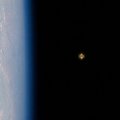 A distant planet appears as a small glowing sphere against the black expanse of space, with Earth's blue atmospheric curve visible on the left edge, evoking the lonely yet awe-inspiring view from orbit that inspired the creation of "Binance Planet."