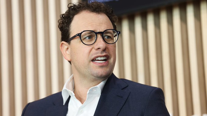A close-up portrait of a man with curly brown hair and dark-rimmed glasses, wearing a white collared shirt and a dark blue suit jacket, speaking against a background of vertical wooden slats.