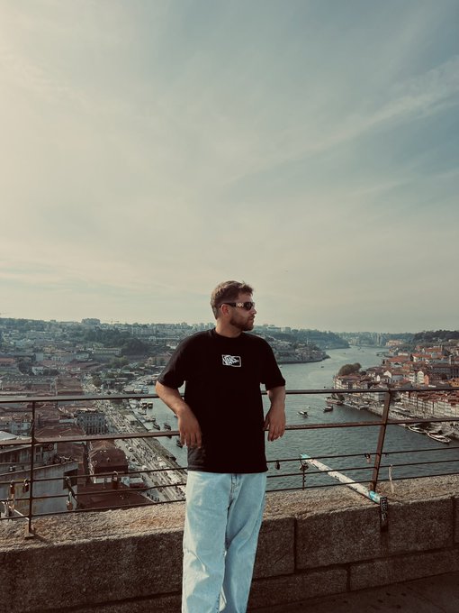 A man wearing sunglasses and a black t-shirt featuring a "N" logo gazes out over a scenic waterfront cityscape, likely Porto or a similar European port, with golden morning light washing over the river and historic rooftops. The image's serene, optimistic vibe pairs well with the post's "Good Morning" greeting, inviting viewers to share their $SOL crypto wallet addresses below.