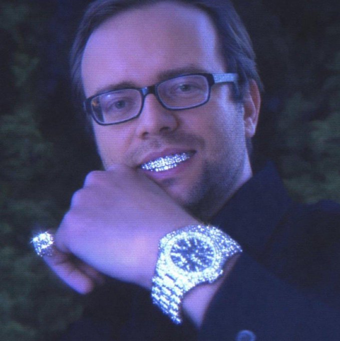 A close-up of a man with glasses smiling and showing off a glittering diamond-encrusted watch and rings against a dark, blue-lit background.