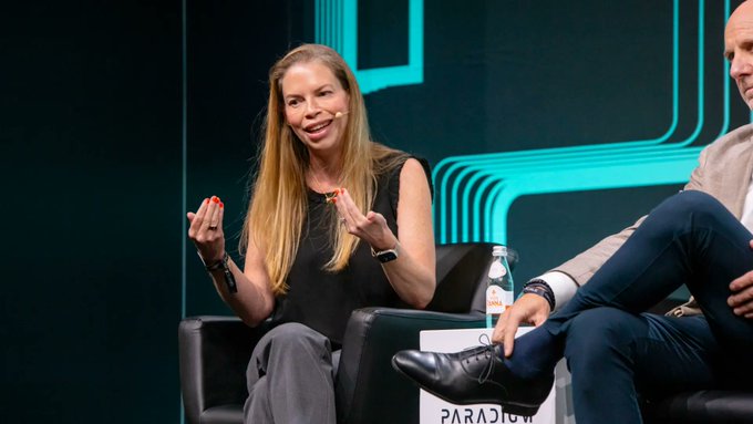 A blonde woman speaks on stage with expressive hand gestures at what appears to be the "Paradigm" event, alongside a man in a light-colored blazer. The image relates to the post as a visual of an industry expert discussing crypto investment strategy, complementing the message about not letting bias cause you to miss market opportunities.