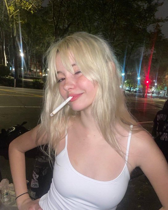 The image features a young woman holding a cigarette, wearing a white tank top, and posing for the camera. She has blonde hair that stands out against the dark background of the photo. The caption "1 night with her or 1 ETH" suggests that she is enjoying herself at an event or gathering where she smoked a cigarette.