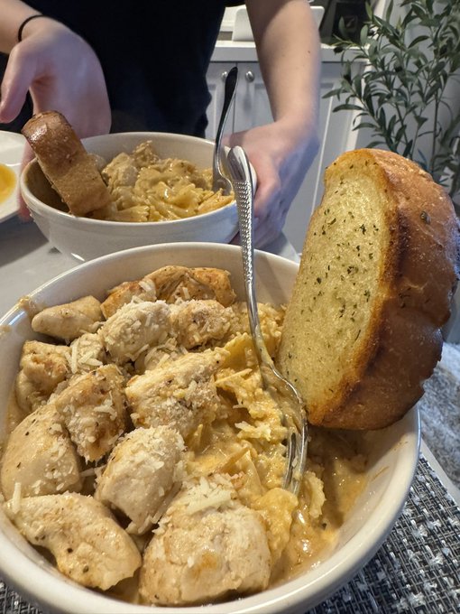 A creamy chicken pasta dish with garlic bread served in white bowls, ready for dinner. The post "dinner is served" perfectly matches the home-cooked meal being presented on the dining table.
