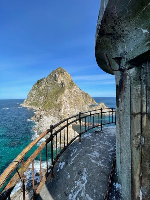 A scenic view from a rusted structure looking out at a rocky island rising from turquoise ocean waters under a clear blue sky.