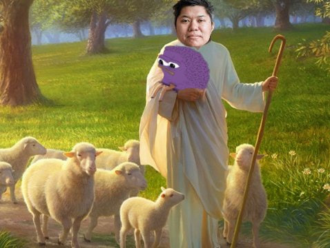 A person dressed as a shepherd in a white robe holds a purple puppet while standing among sheep in a green meadow with trees.