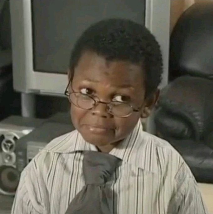 A young boy wearing glasses, a striped shirt, and a gray tie makes a skeptical or unimpressed facial expression in what appears to be a living room or office setting.