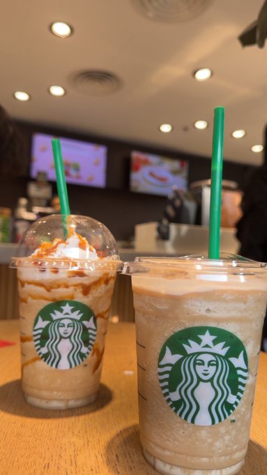 Two Starbucks iced beverages in clear plastic cups with green straws sit on a wooden table, with the left cup containing a caramel-drizzled frappuccino topped with whipped cream and the right cup holding a plain iced coffee, in what appears to be a cafe setting.